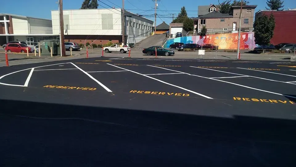 Newly paved parking lot with reserved spaces, cars, and buildings in the background.