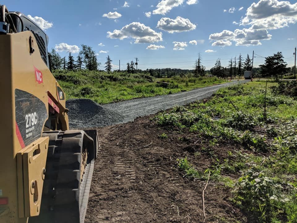 Yellow skid steer compacting gravel road in a field under a blue sky with clouds.