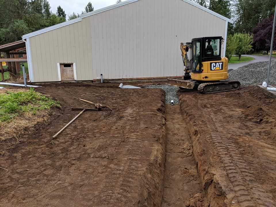 Construction site with a small excavator near a light tan building. A trench is being dug in the dirt.