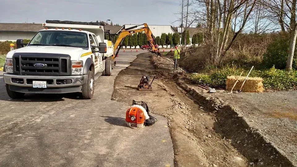 Construction site with excavator and truck; workers digging and blowing debris.