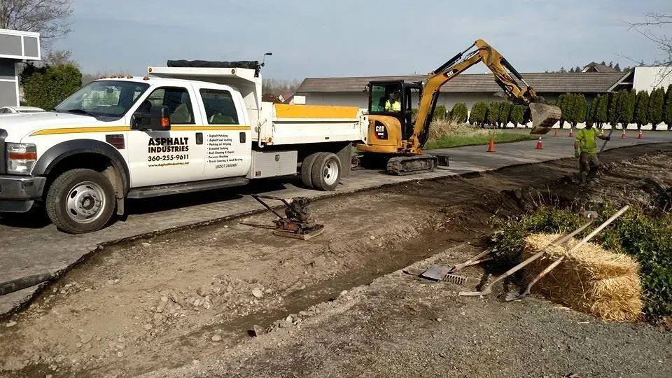 A white work truck and small excavator digging a trench on a paved area. A bale of hay sits nearby.