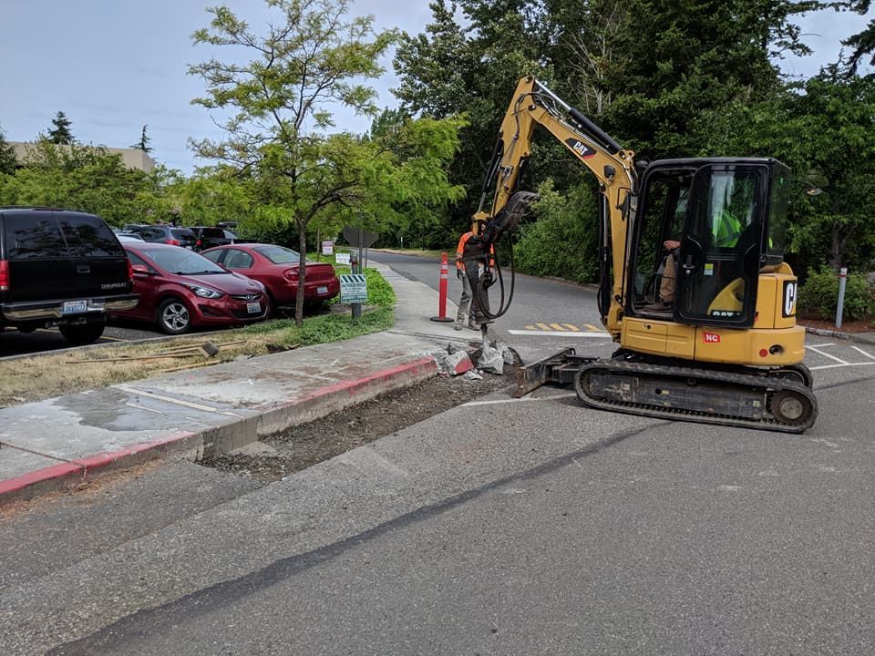 Yellow excavator working on a sidewalk next to a road and parked cars.