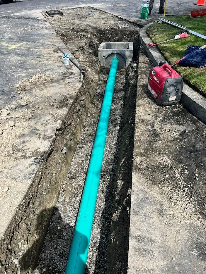 Construction site with green pipe in a trench. A concrete box is attached. A generator and curb are visible.