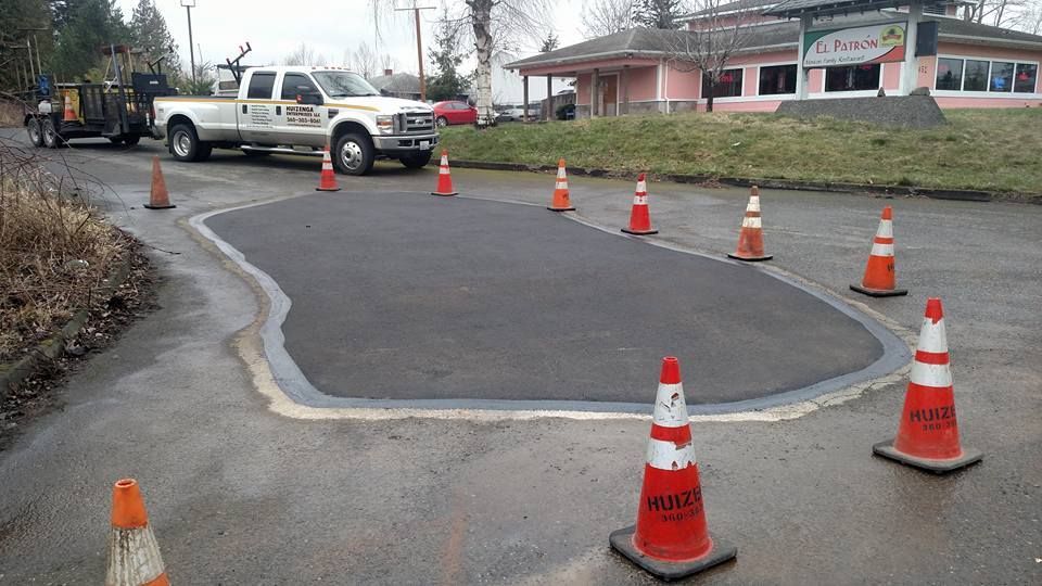 Asphalt patch repair on a road, orange cones marking perimeter, truck and trailer in the background.