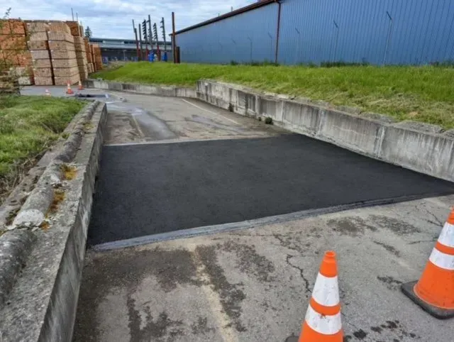 Asphalt repair in a concrete-lined road section, orange traffic cones, and industrial building in the background.