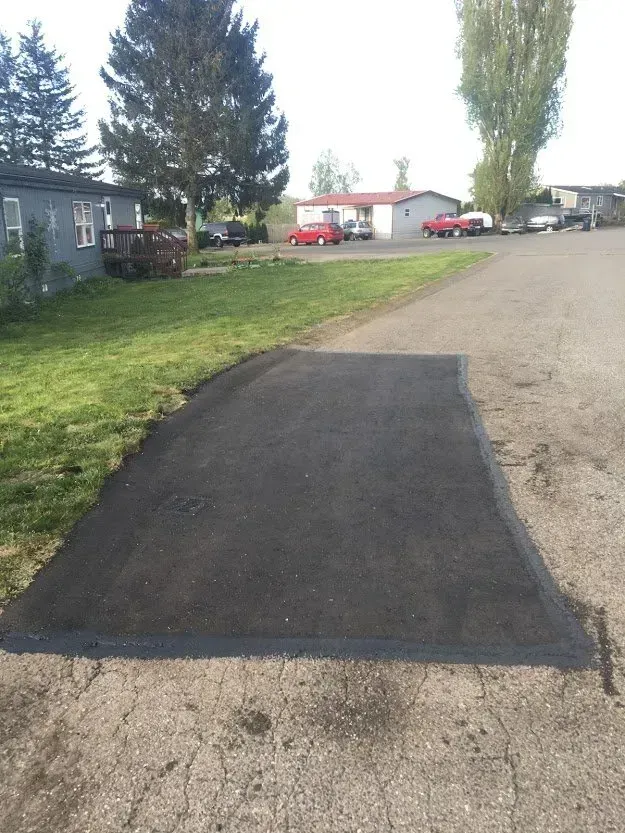 Newly paved asphalt section on a road, merging with the older, weathered pavement. Buildings and cars in the background.