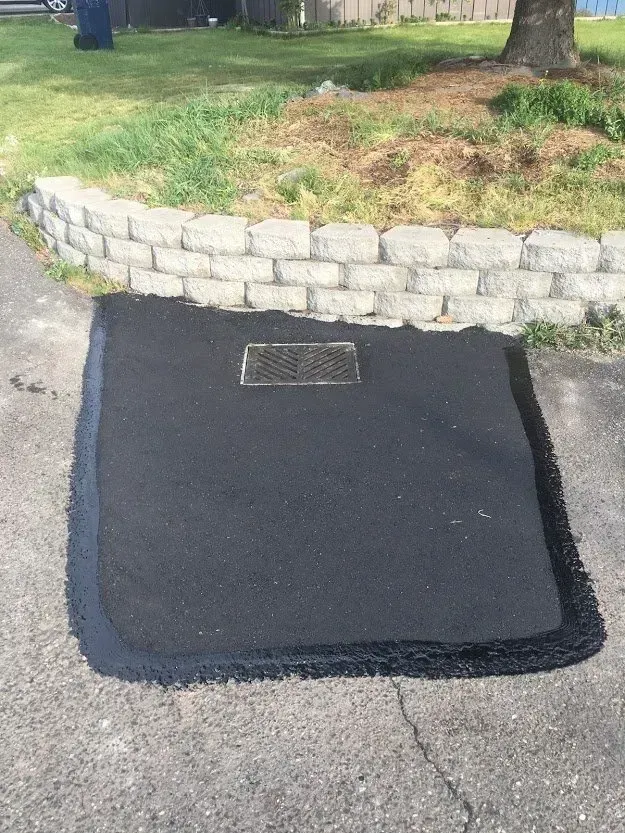 Fresh asphalt patch with a sewer grate on a driveway, bordered by a brick wall and grass.