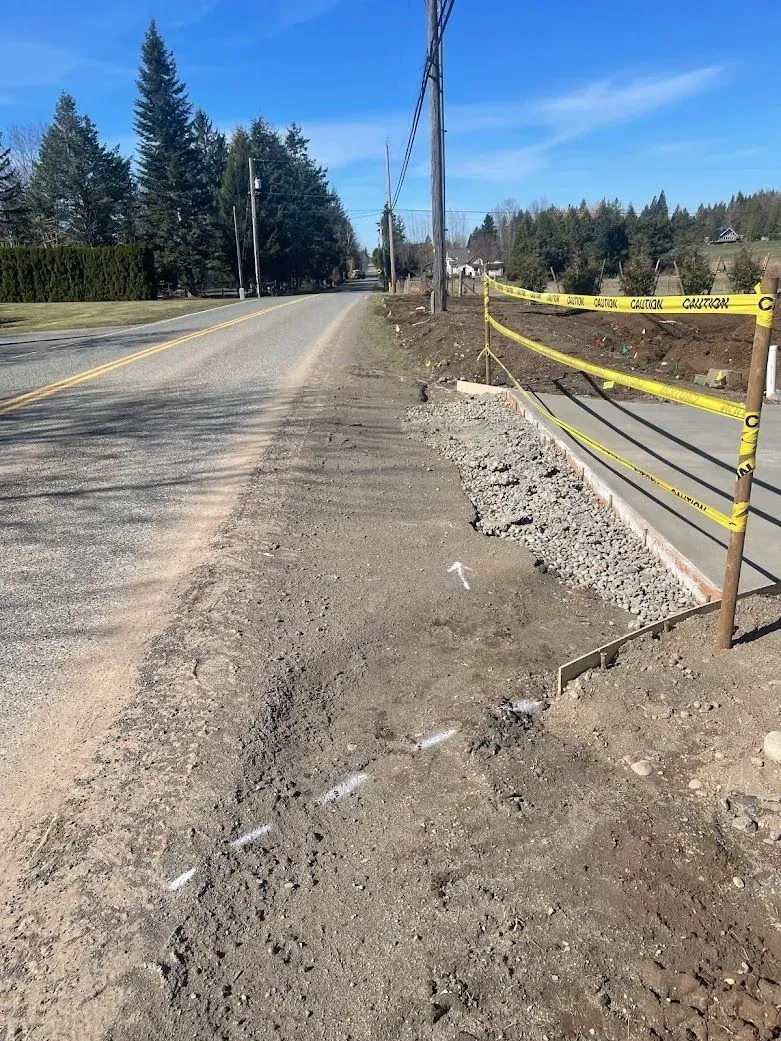 Roadside construction site with caution tape. Paved road, dirt path, and concrete edging are visible. Blue sky.
