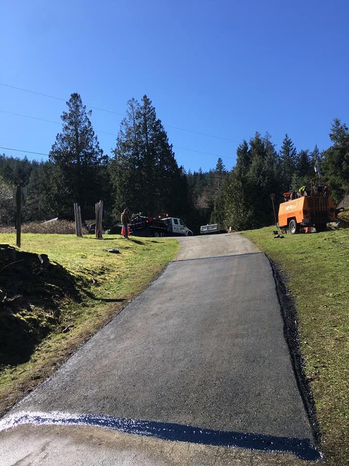 Paved road leading uphill through grassy area, with trees in the background and work vehicles present.