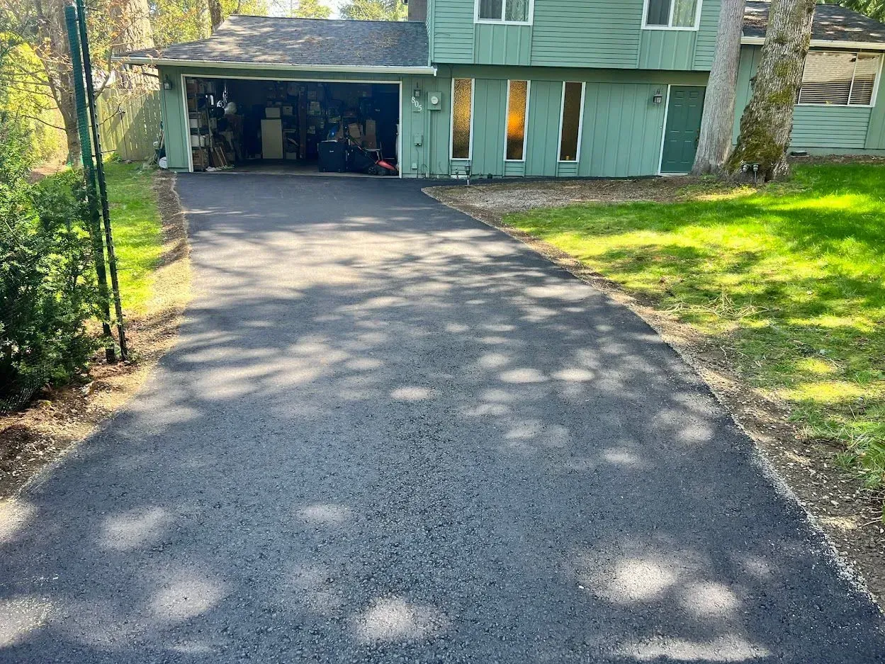 Asphalt driveway leading to a green house with an open garage, surrounded by grass and trees.