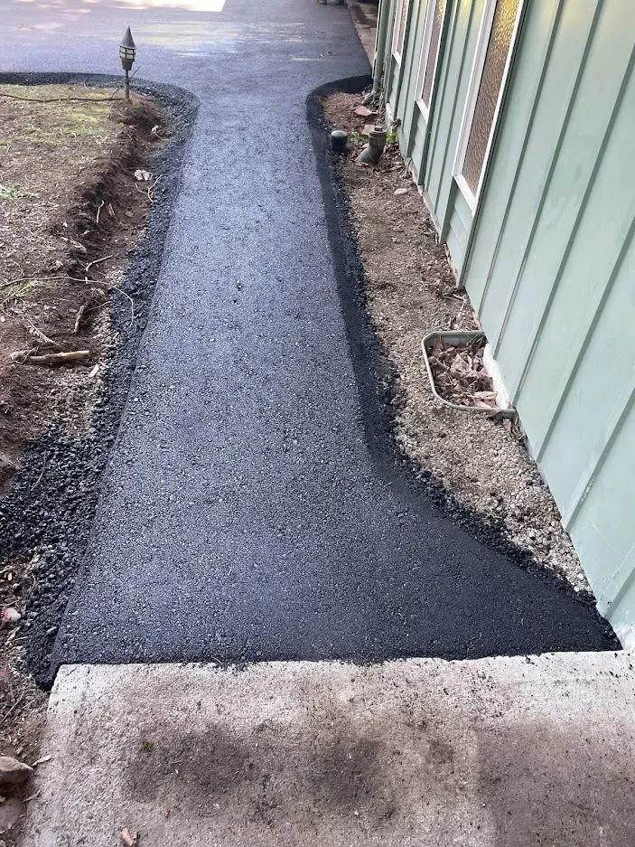 Asphalt pathway leading from concrete steps, bordered by dirt and a building with green siding.