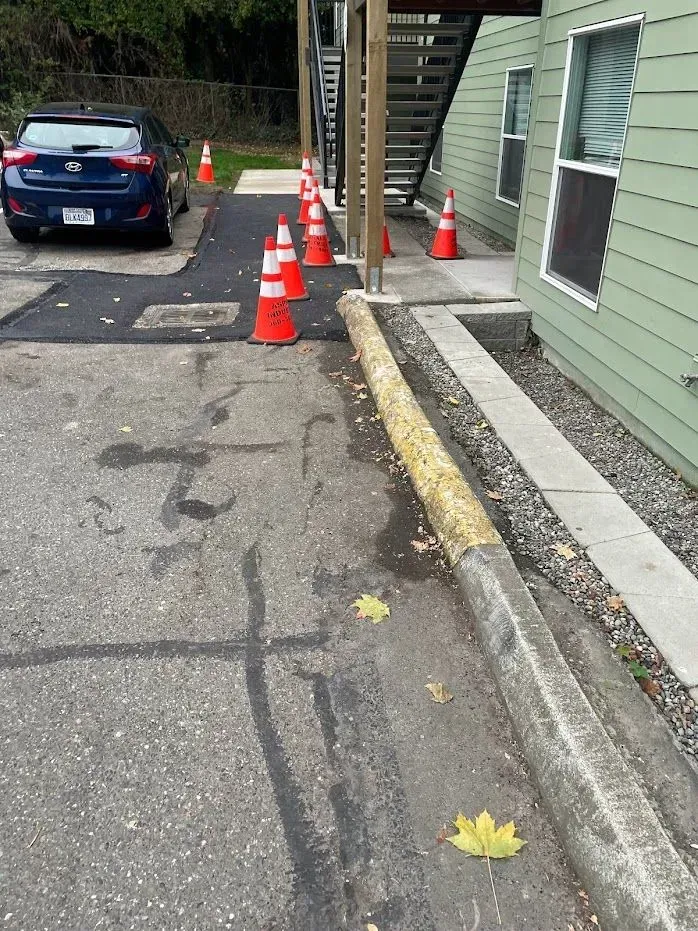 Asphalt repair area marked with orange traffic cones, next to building with stairs. Blue car is parked.