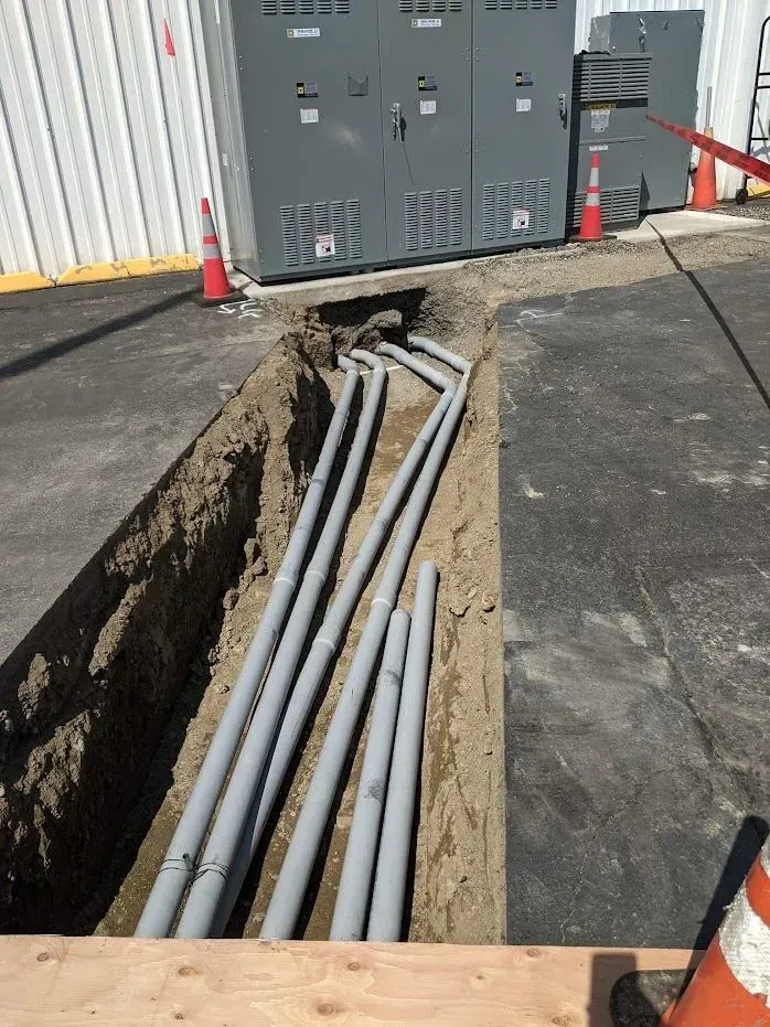 Trench with gray pipes, near electrical boxes and cones, outside.