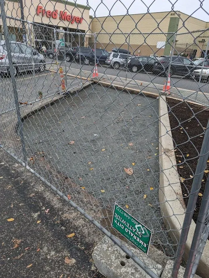 Fenced-off area with gravel and soil, next to a Fred Meyer store parking lot.