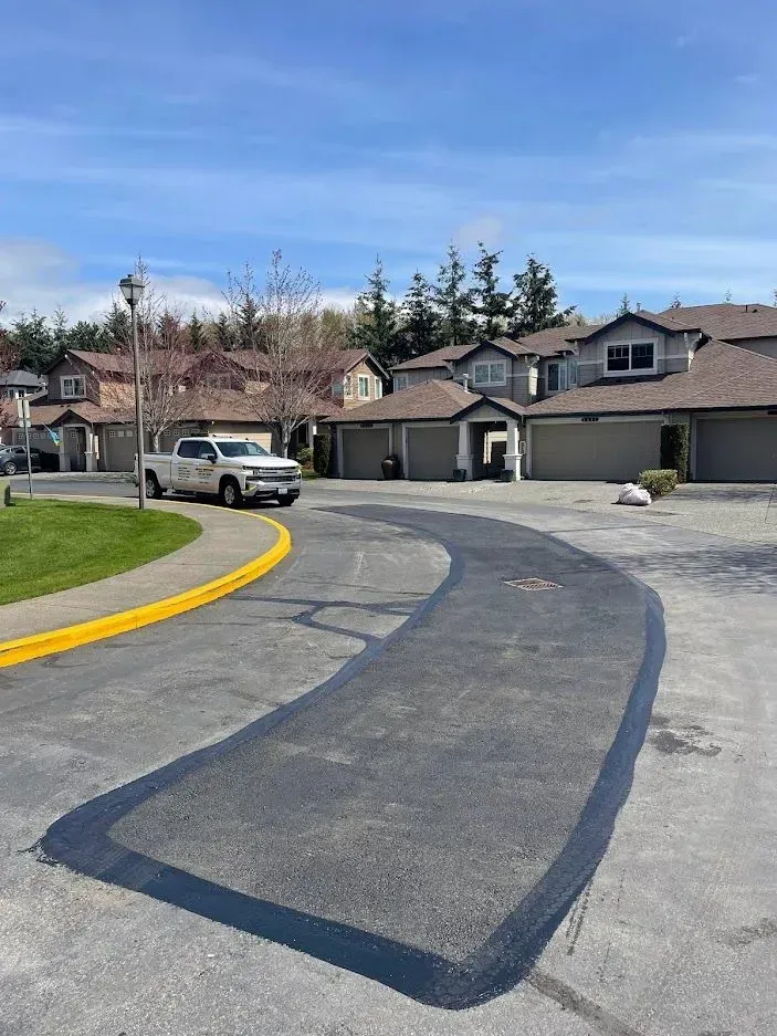 Newly paved asphalt section on a residential street; a truck is parked nearby, houses and blue sky background.