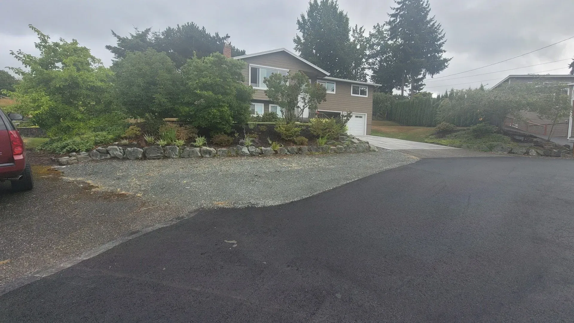 A house with a gravel driveway, surrounded by trees and bushes, on a cloudy day.