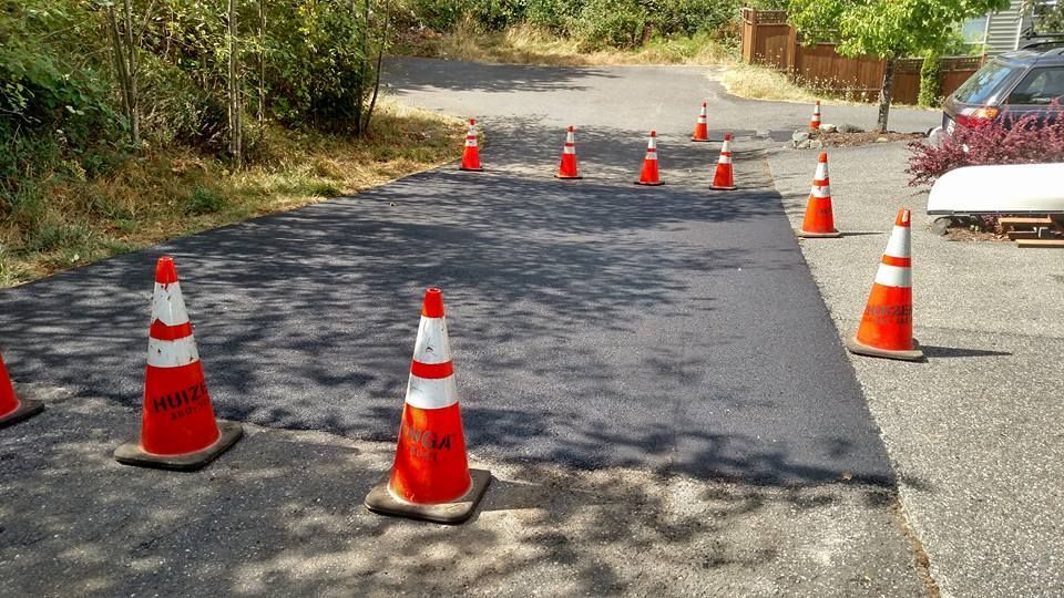 Orange cones line a freshly paved asphalt driveway; trees and a car are in the background.