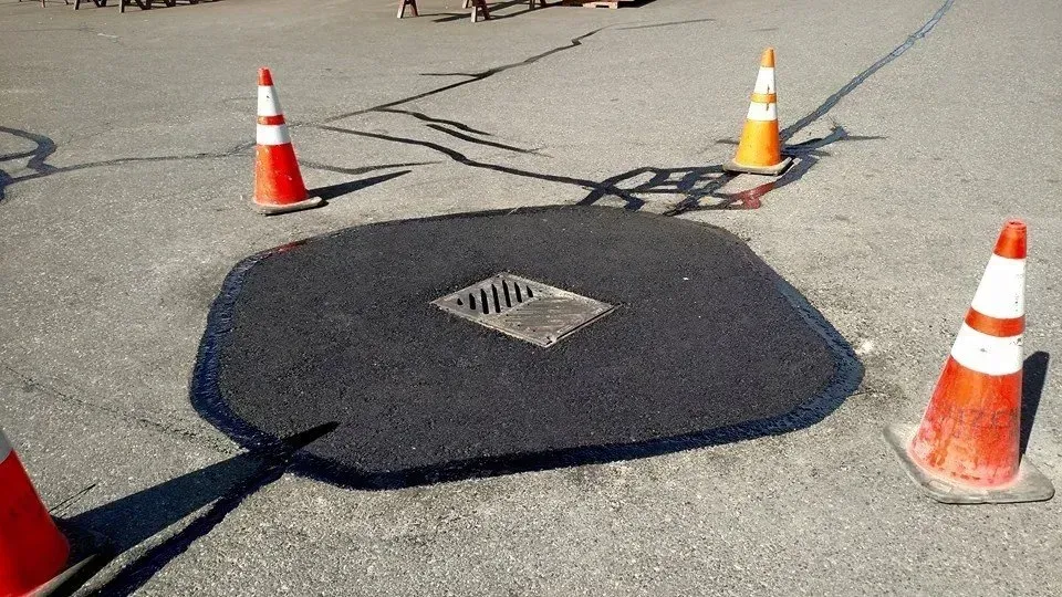 Asphalt patch with drain, surrounded by orange and white traffic cones on a paved surface.