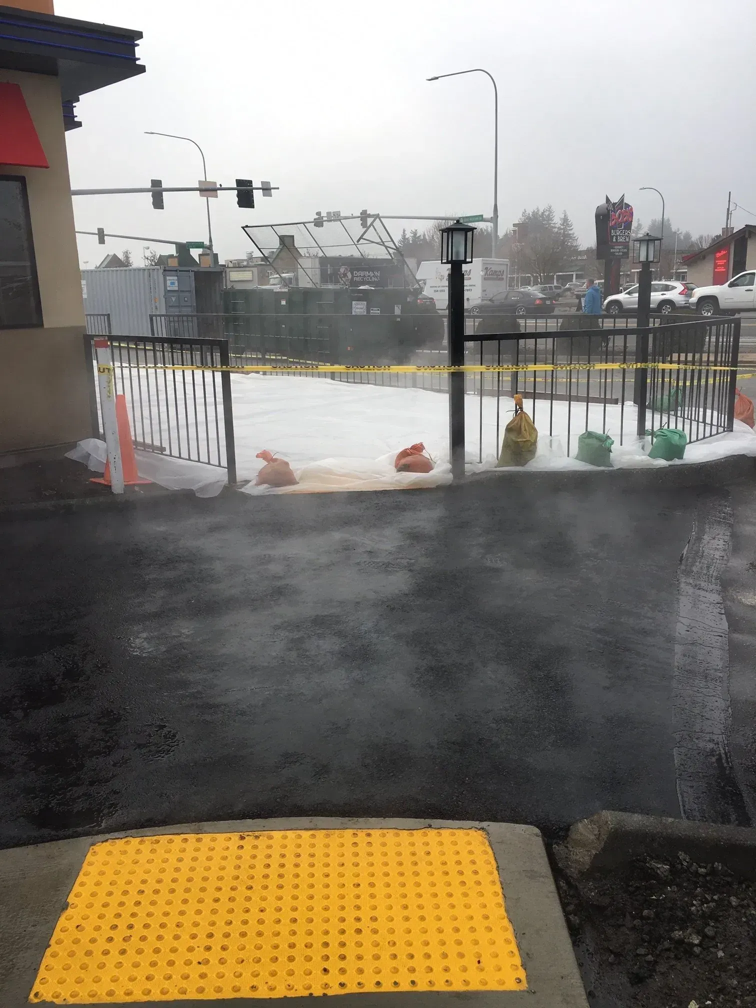 Exterior view of a building with a fenced-off area covered in white substance. Yellow caution tape and traffic cones visible.
