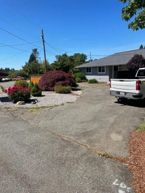 Asphalt driveway in front of a gray house with a parked white pickup truck, shrubbery, and a clear blue sky.