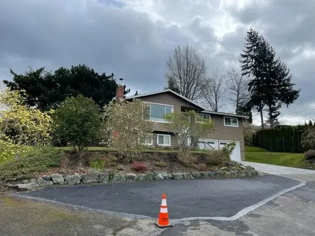 House with two-car garage, on a slight hill, gray siding, dark asphalt driveway, cloudy sky.