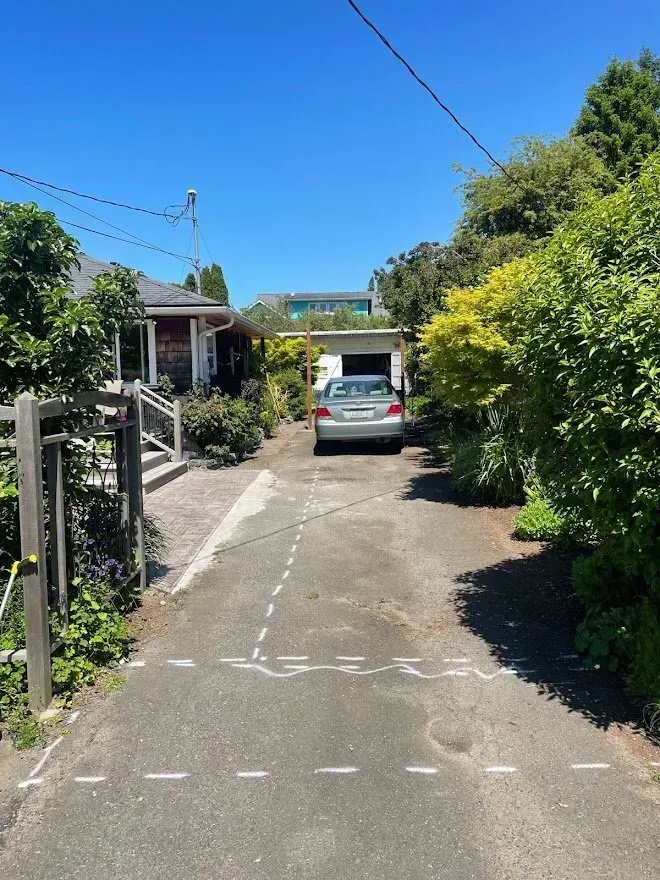 Asphalt driveway with a car parked in front of a garage, surrounded by houses and greenery on a sunny day.