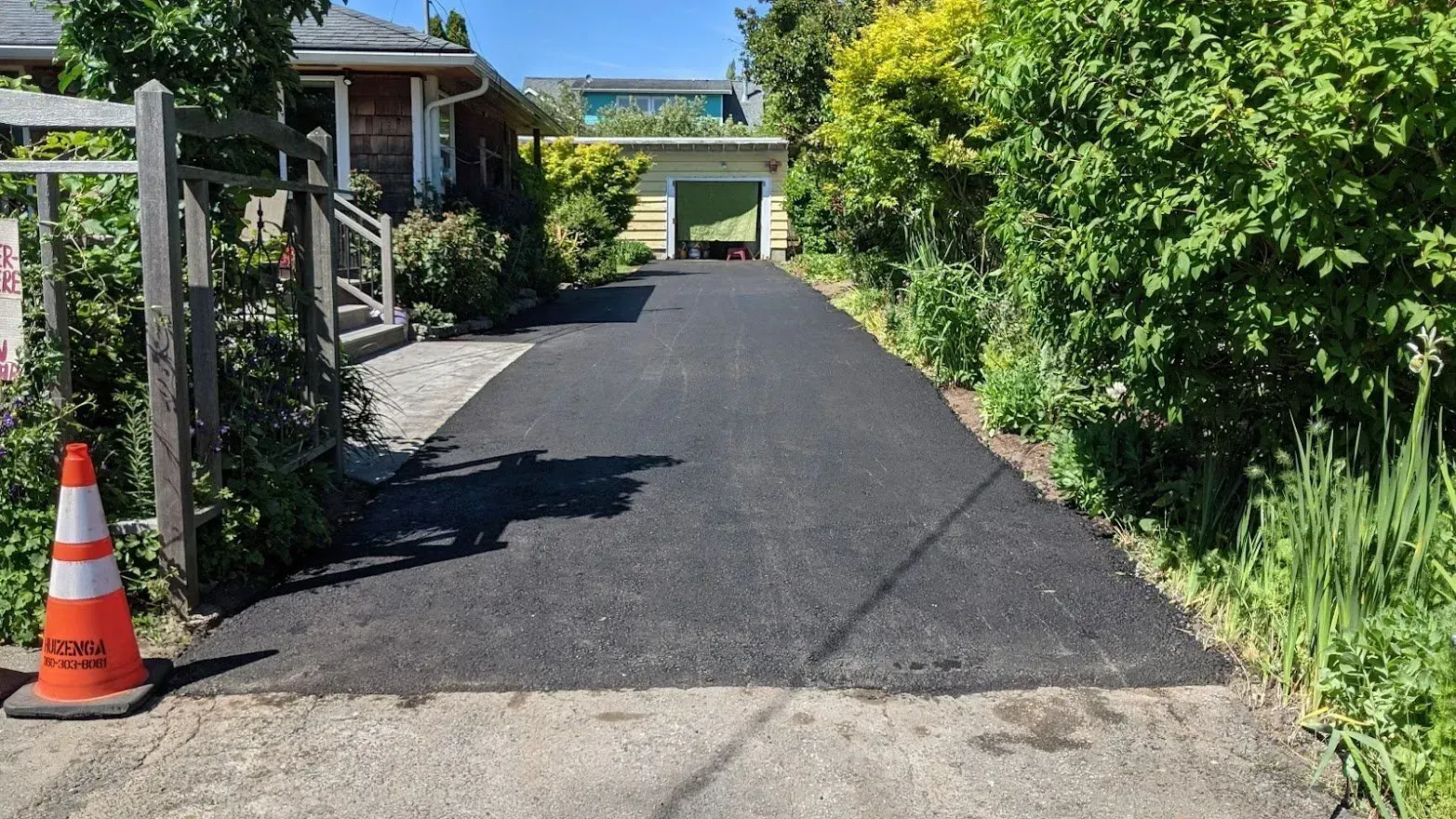 Newly paved asphalt driveway leading to a garage, flanked by greenery and a house. A traffic cone is on the left.