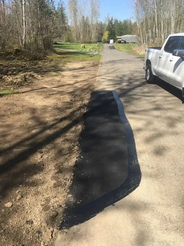 Asphalt road repair; dark asphalt patch next to dirt shoulder; white truck on the right.