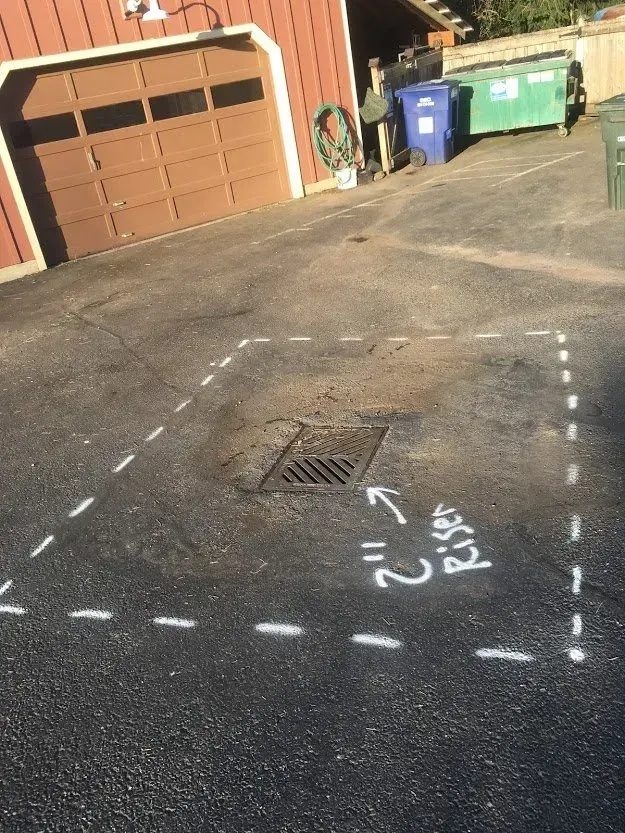 Asphalt driveway with drain marked for a 2-inch rise with dashed white lines; garage and trash bins in the background.