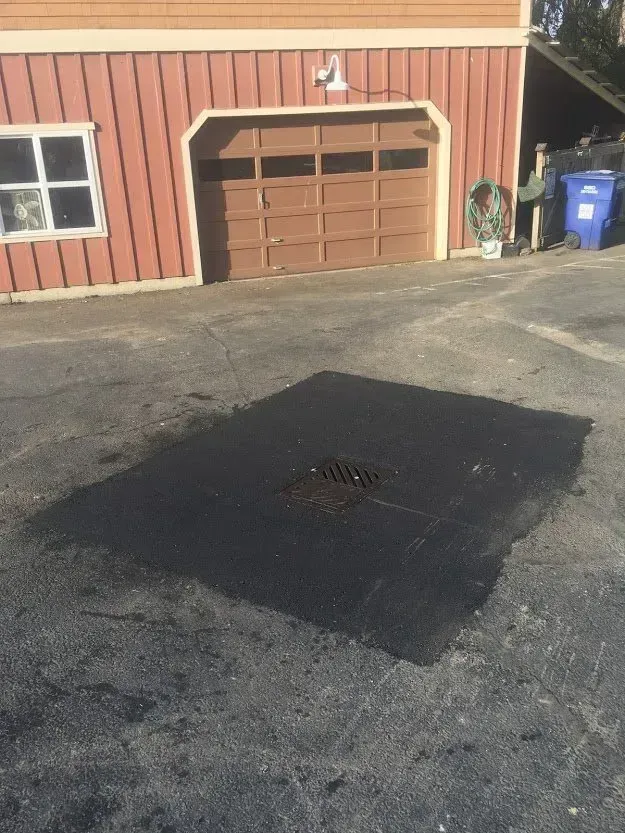 Patch of fresh asphalt covers a drain in a paved driveway near a garage with a brown door.