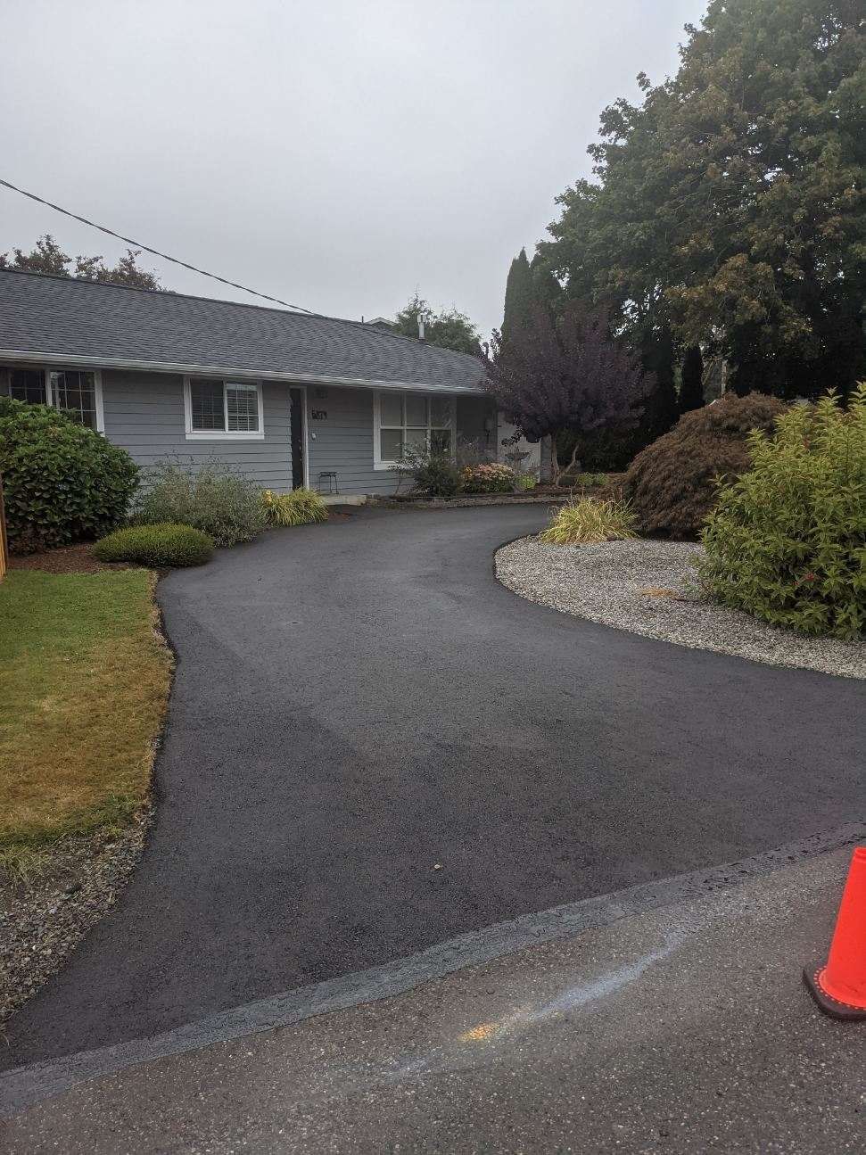 Newly paved asphalt driveway leading to a gray house with a small front yard and landscaping.
