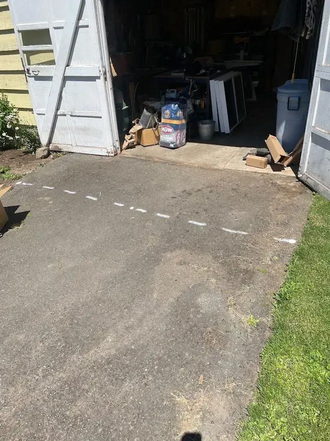 Asphalt driveway with white dashed lines leading to an open garage filled with clutter.