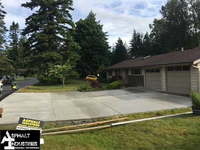 Newly paved concrete driveway in front of a house, green lawn, trees, and cloudy sky.