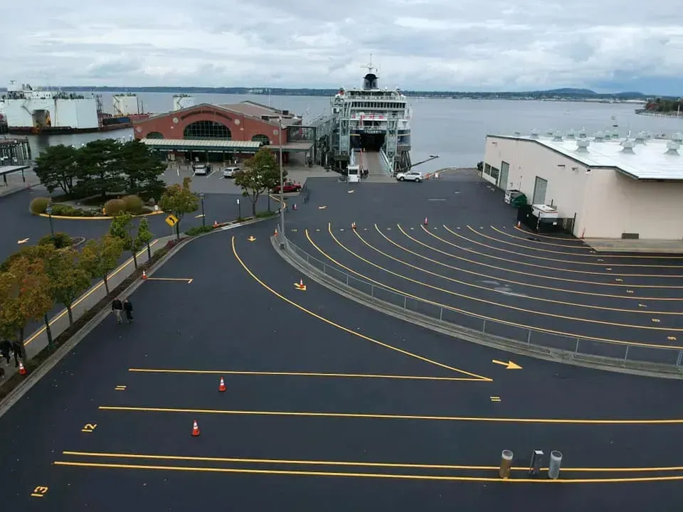 Ferry docked at a terminal. Asphalt road with lane markings. Buildings and water in the background. Overcast sky.