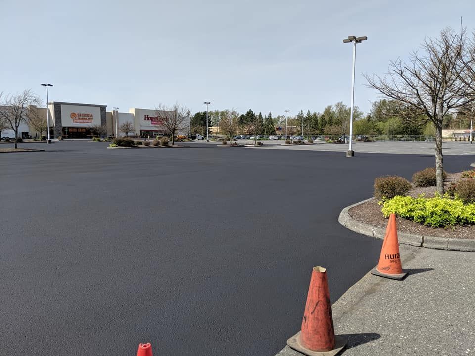 Newly paved, dark asphalt parking lot with orange cones, in front of retail stores under a clear sky.