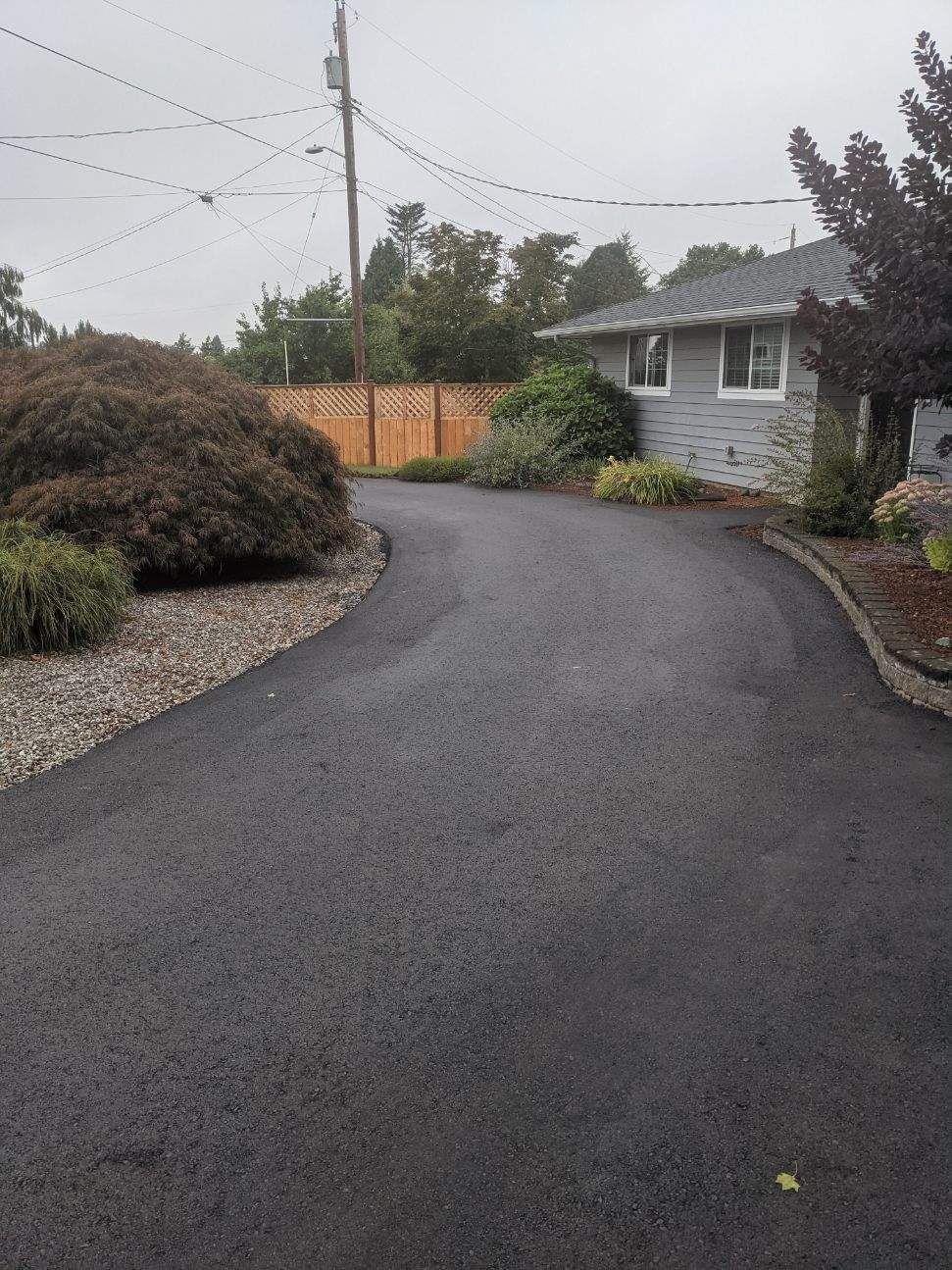 Asphalt driveway curves toward a gray house with a wooden fence in the background under an overcast sky.