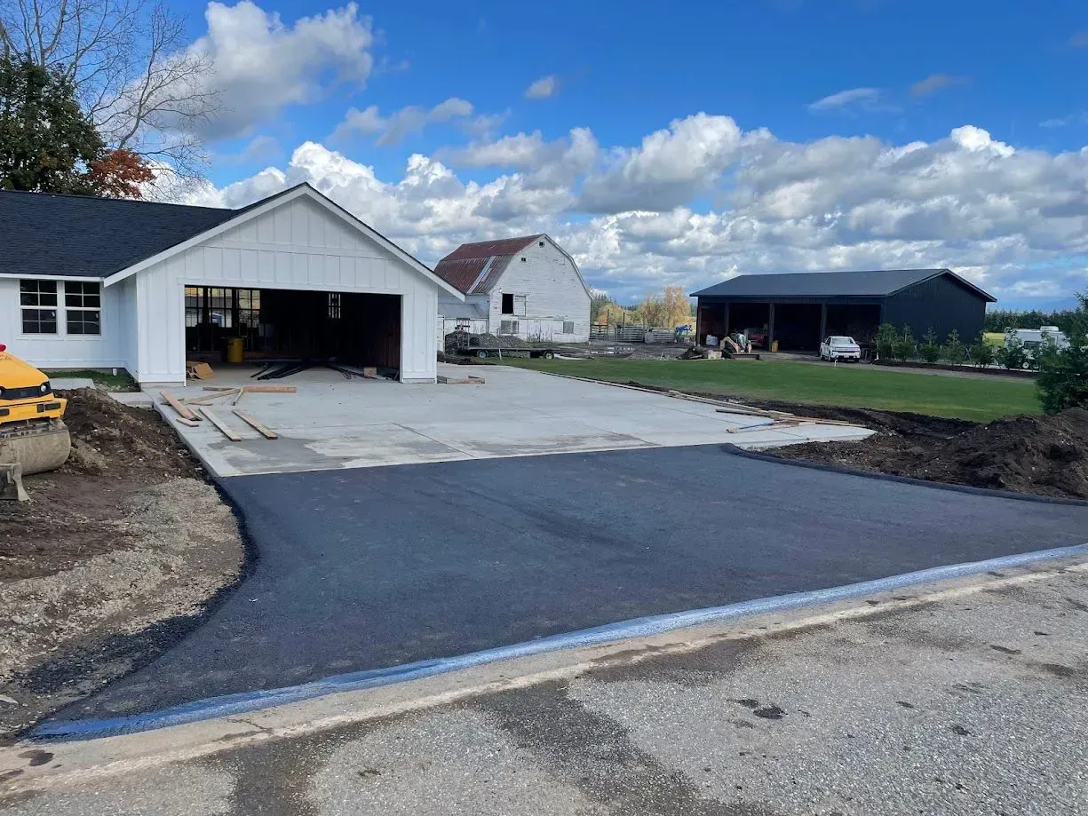 Asphalt driveway leading to a white garage, next to a house and barn, under a cloudy sky.