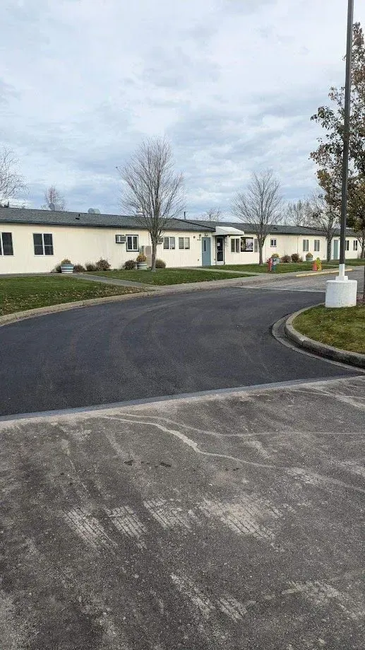 Paved road curves towards a light-colored, single-story building under a cloudy sky.