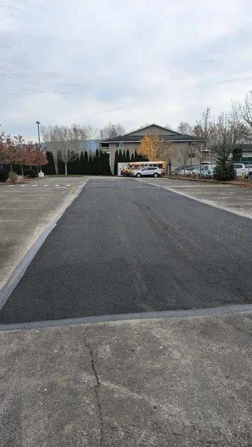 Newly paved asphalt area in a parking lot, with a small bus and a building in the background.
