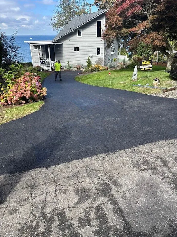Asphalt driveway leads to a gray house by a body of water. A person in a high-vis vest stands on the driveway.