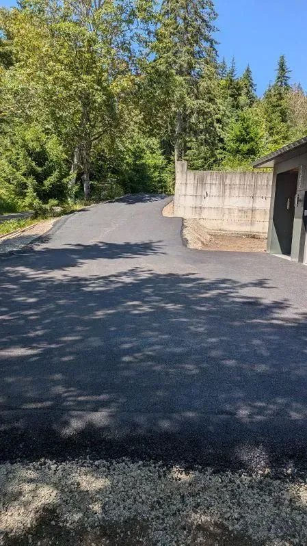 Paved driveway curves uphill past a concrete wall and building, trees in background.