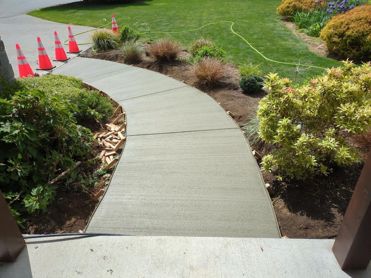 Curved concrete sidewalk bordered by shrubs and grass, with orange traffic cones marking the left edge