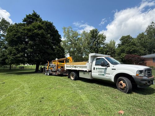 White utility truck towing yellow equipment trailer across a grassy field under a sunny blue sky