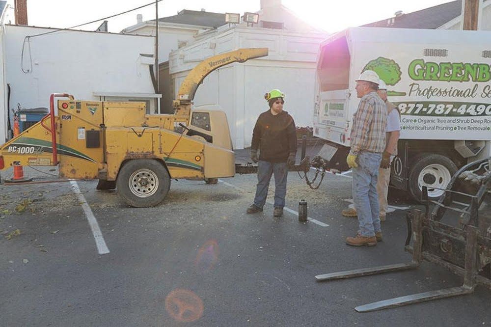 Workers beside a yellow wood chipper and truck on a sunny street, with debris and hoses on the ground