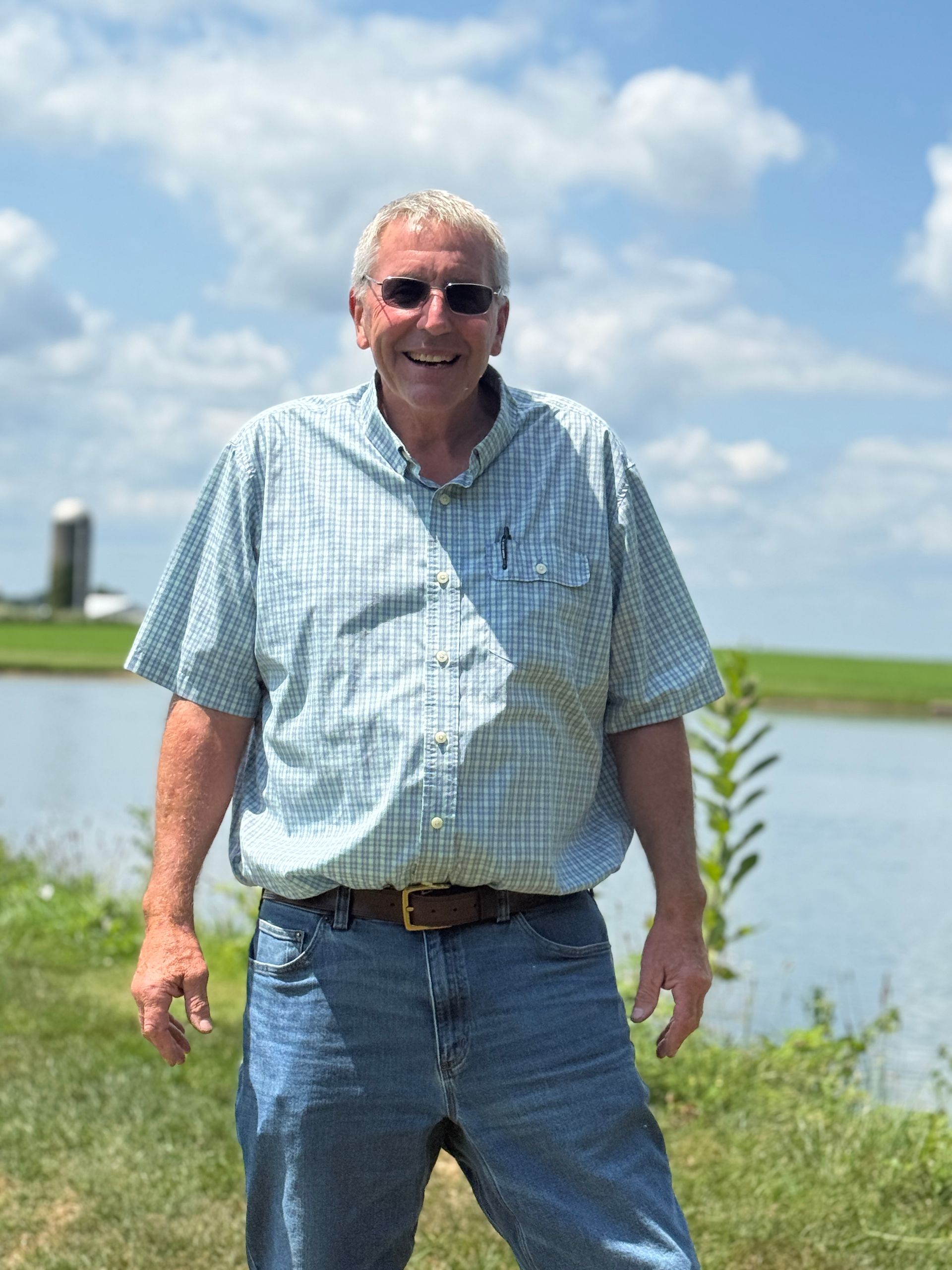 Man standing outdoors by a waterfront on a sunny day, wearing a light blue shirt and jeans.