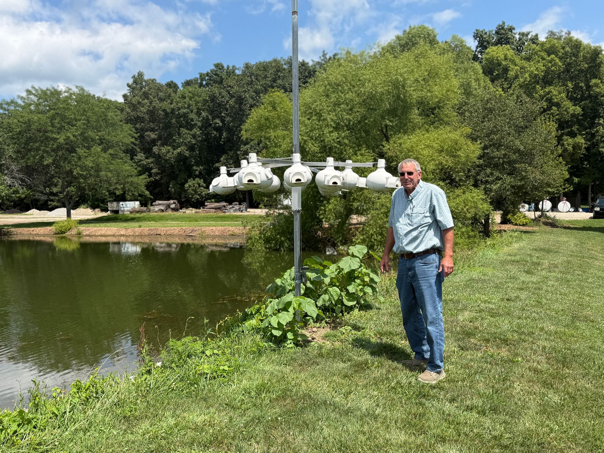 Man standing by a pond in a green park beside a white lamp post under a blue sky