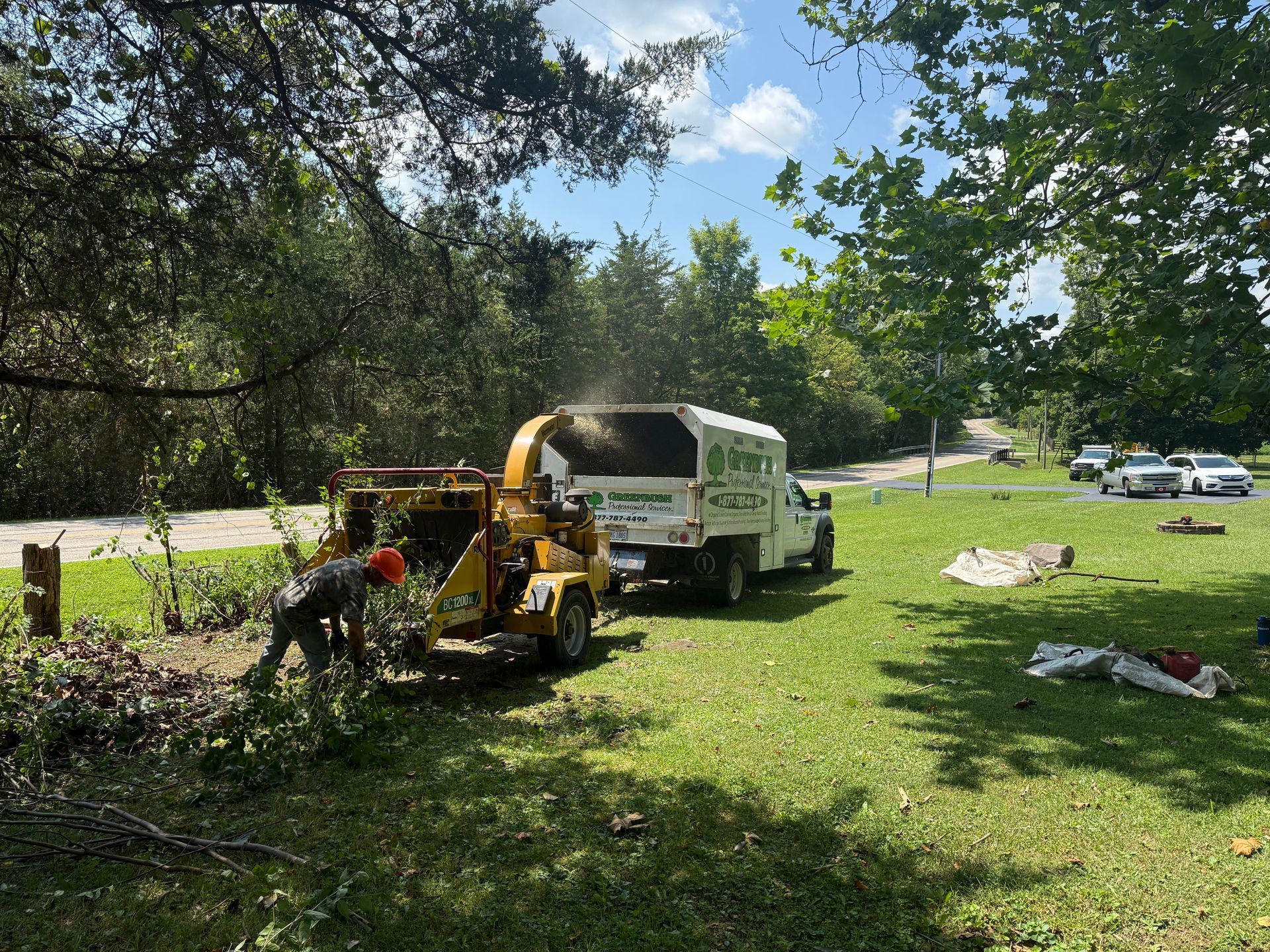 Workers clearing brush with a wood chipper beside a white truck in a grassy, wooded park area.