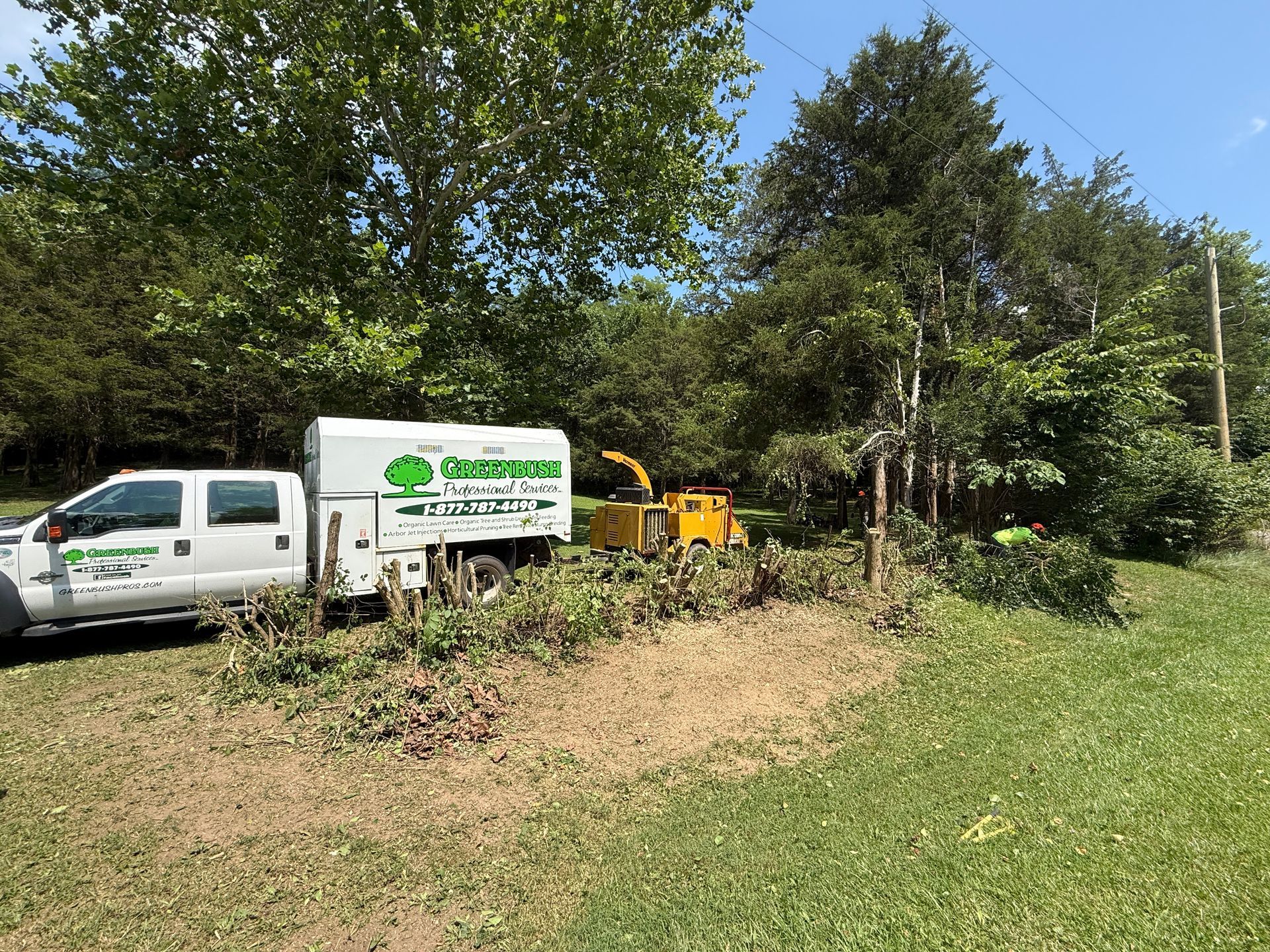 White truck and yellow utility machine beside a grassy field and trees under a clear blue sky