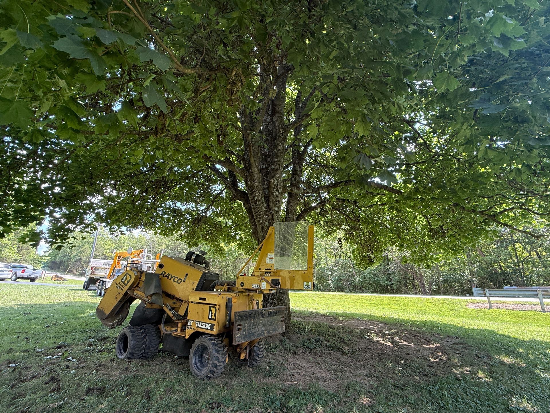 Yellow utility machine parked under a large tree in a grassy field, with a road and parked cars in the background