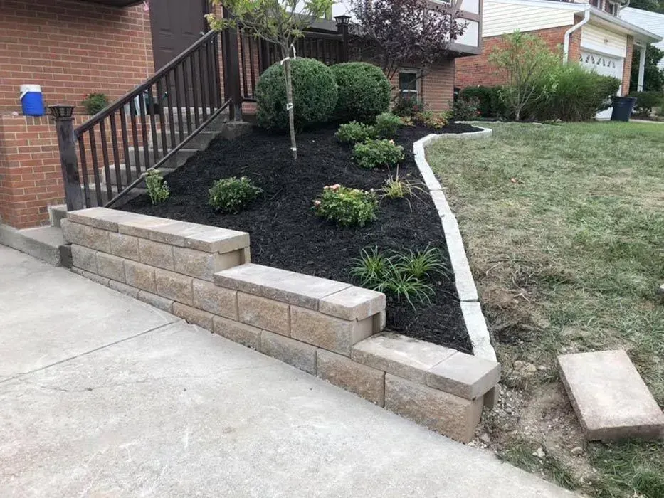 Landscaped front yard with brick retaining wall, black mulch, shrubs, and concrete steps beside a driveway